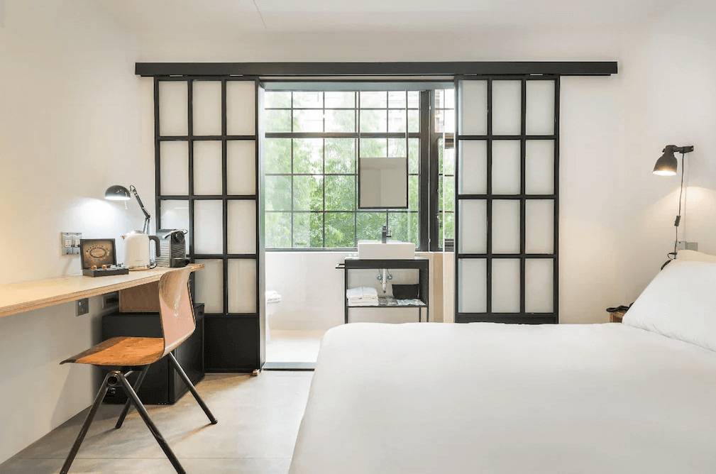 【西門町住宿】艾特文旅的雙人房，搭配大面窗戶引入自然光。房內擺放一張雙人床，牆面裝有黑色金屬框架的日式拉門，將浴室與房間分隔開來。洗手台設於窗前，提供通透開闊的視野，簡約的木質書桌與鐵椅增添復古氛圍。