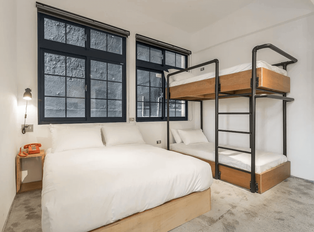 【西門町住宿】艾特文旅的家庭房，設有一張雙人床與一組黑色金屬框架的上下舖，適合多人入住。房間以簡約白色牆面搭配深色窗框，營造出工業風設計感。床邊擺放復古橙色電話，增加懷舊元素。