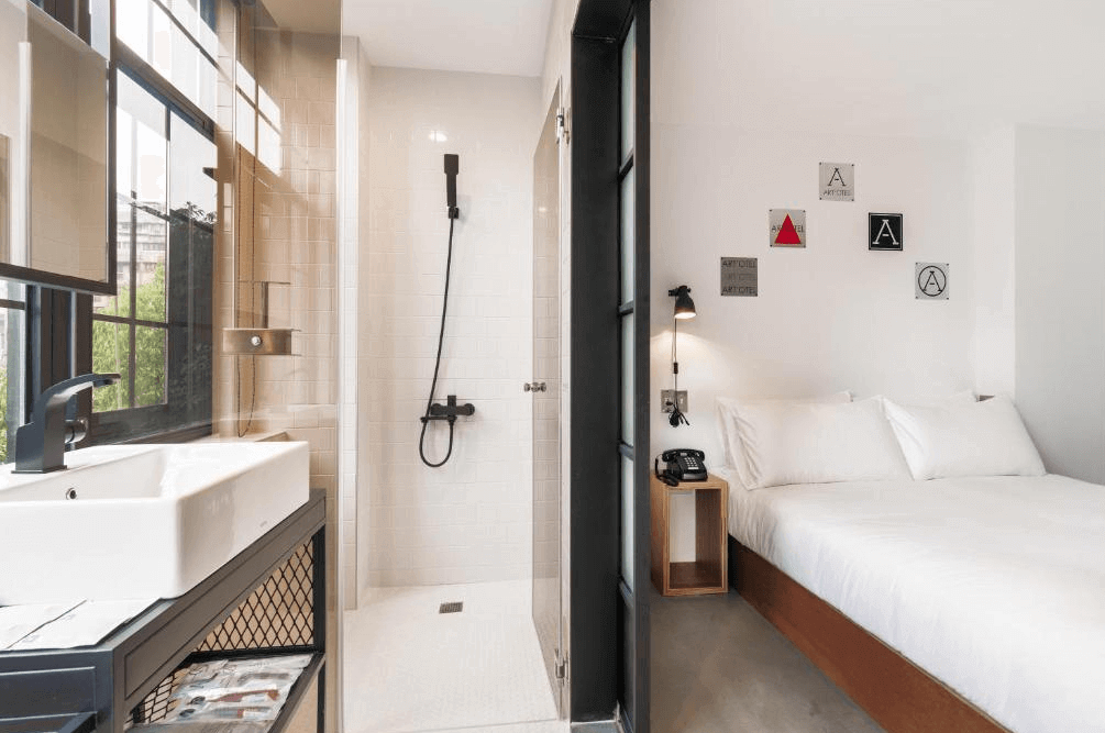 【西門町住宿】艾特文旅半開放式浴室設計，採用白色磁磚與黑色五金配件，乾淨俐落。洗手台位於窗邊，搭配黑色金屬框鏡面，浴室內設有獨立淋浴間。房內擺放雙人床，牆面裝飾幾何圖案，營造現代感。
