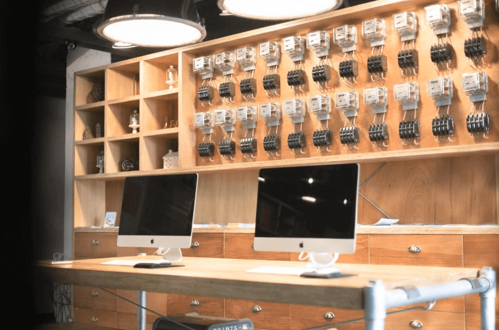 【西門町住宿】艾特文旅的公共空間，擺放兩台蘋果電腦供住客使用。背景牆上排列著多個老式電錶，搭配木質櫃體與金屬燈具，展現獨特復古工業風。開放式書架內擺放各式裝飾品，增添藝術氣息。