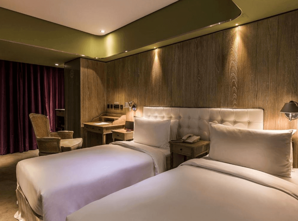 【西門町住宿】薆悅酒店西寧館的雙床房設計，兩張單人床鋪著純白色床單，搭配柔軟的米白色皮革床頭板。牆面採用木質裝飾，搭配暖色間接燈光營造溫馨氛圍。房內設有書桌、藤椅與深紫色窗簾，增添復古質感。