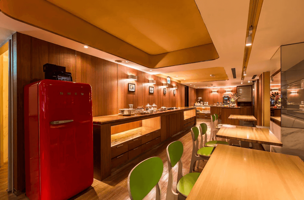 【西門町住宿】薆悅酒店西寧館的自助餐廳空間，木質裝潢搭配暖黃色天花板燈光，營造舒適的用餐氛圍。餐檯上擺放各式早餐選擇，並配有復古風格的紅色SMEG冰箱，桌椅選用淺木色搭配綠色椅背，展現活潑氣息。