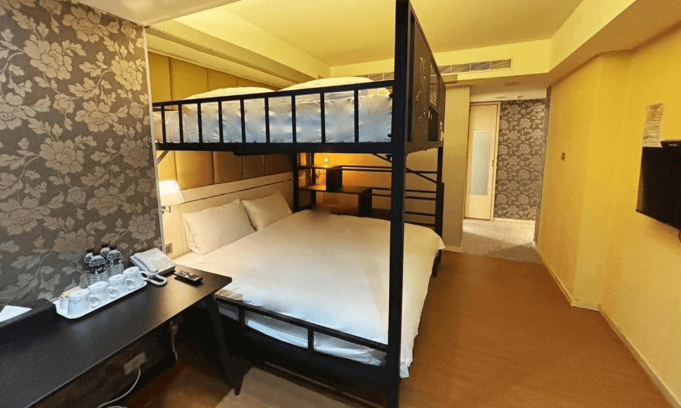 【松山機場住宿】「豪城商旅民生館」的家庭房，特色為上下舖床型設計，下鋪為雙人床，上鋪為單人床，適合親子或多人入住。房間內佈置溫馨，牆面帶有花卉壁紙，並配備書桌、茶具與瓶裝水。