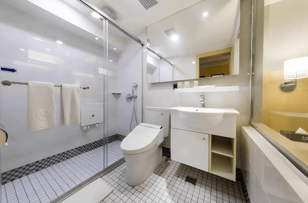 【松山機場住宿】「豪城商旅民生館」的浴室空間，採用明亮的白色系設計，提供乾溼分離的淋浴間與現代化馬桶。洗手台下方設有收納空間，並提供基本備品，整體乾淨且實用。