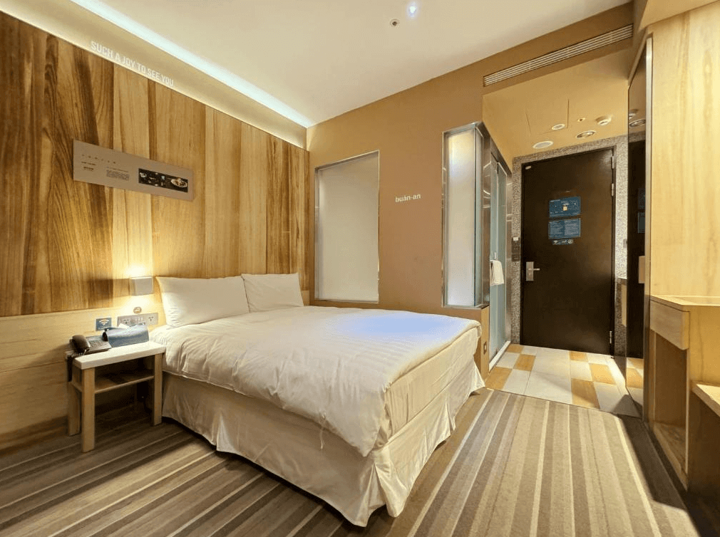 【台北親子飯店】路境行旅的溫馨客房，以木質裝潢營造自然氛圍。房內設有一張舒適的雙人床，床頭牆面以木紋裝飾，搭配柔和燈光，營造放鬆的住宿體驗。房門旁的玻璃浴室設計增添現代感。