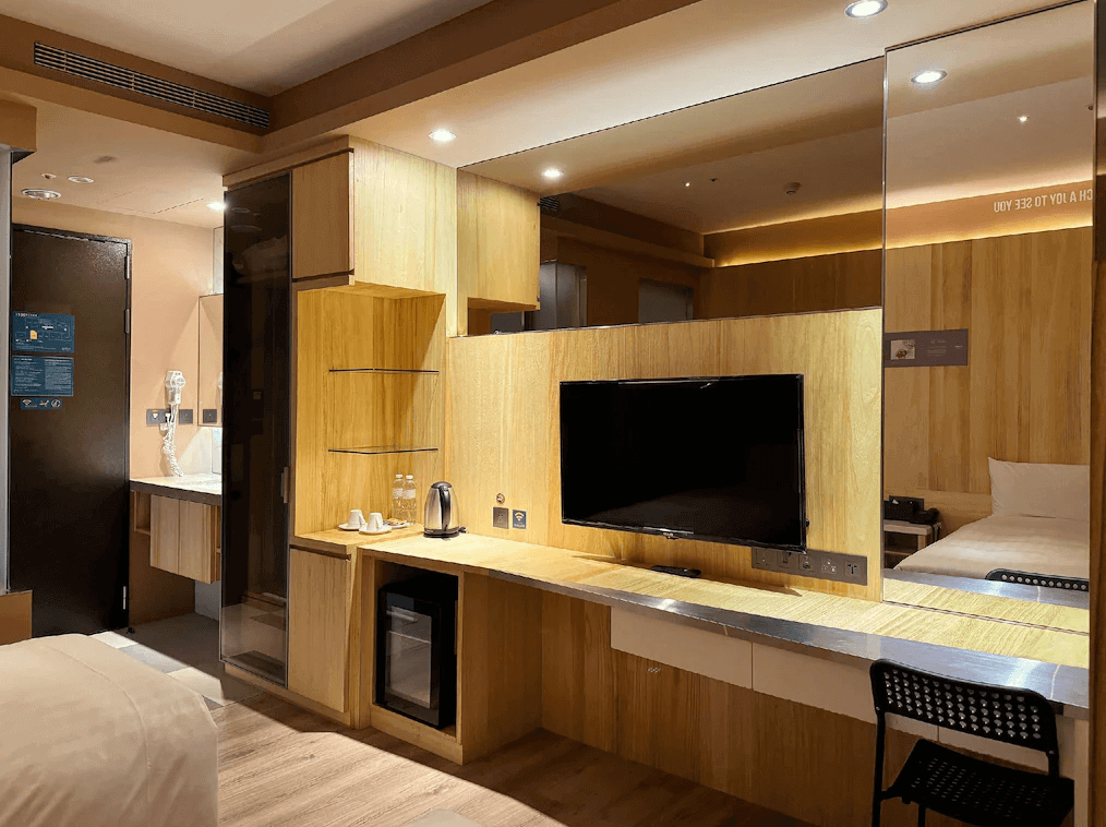 【台北親子飯店】路境行旅客房內的多功能設計，包括木質書桌、內嵌電視、大型鏡面及開放式收納空間。房內提供熱水壺、瓶裝水和迷你冰箱，展現實用與設計感兼具的住宿環境。