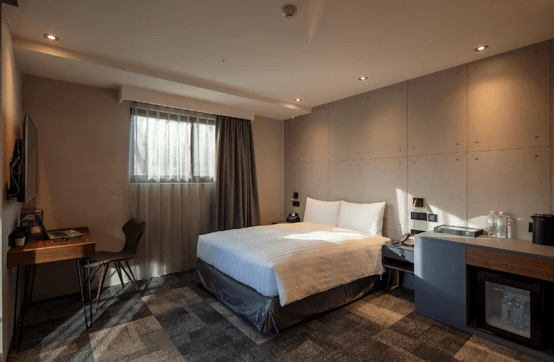 【台北車站住宿】路徒Plus行旅主題館標準雙人房，採用工業風設計，搭配灰色水泥牆與溫暖的間接照明。房內設有一張大床、辦公桌、電視和迷你吧，窗戶提供自然採光，營造簡約現代感。
