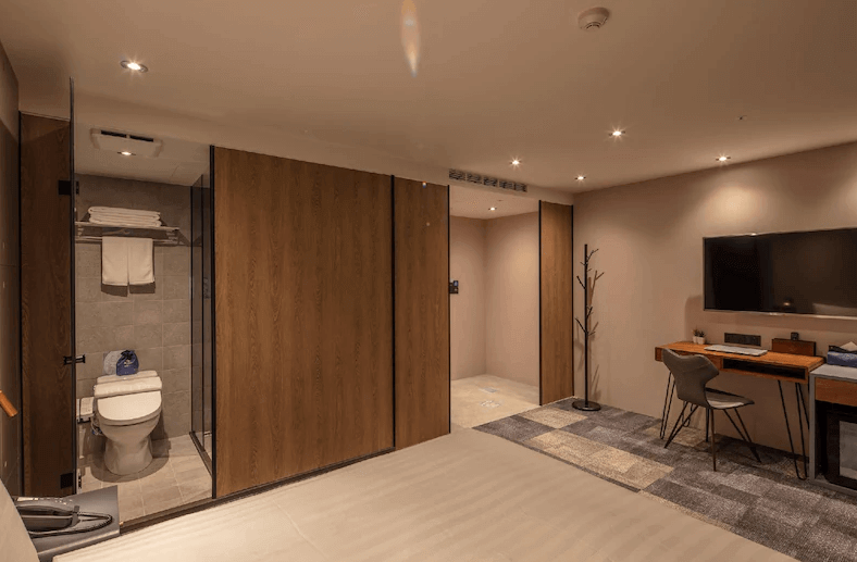 【台北車站住宿】路徒Plus行旅 主題館的開放式工業風客房，內部配備大床、辦公桌與壁掛式電視。浴室區域採玻璃與木質門設計，空間規劃簡約而富有設計感，並融合柔和燈光提升溫馨氛圍。