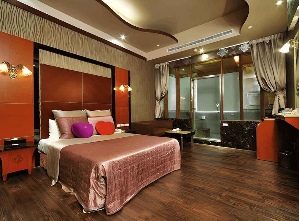 【金山溫泉】金湧泉SPA溫泉會館的溫泉套房，搭配深色木質地板與紅棕色牆面設計。大床上擺放著心形抱枕，展現浪漫氛圍。房間內部設有寬敞的休憩區與大型玻璃浴室，適合情侶或夫妻入住，享受舒適的度假體驗。