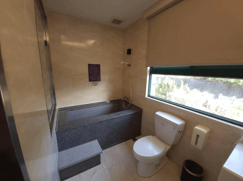 【金山溫泉】金湧泉SPA溫泉會館的溫泉浴室內景，設有深色石材打造的泡湯池與現代化衛浴設備。大片窗戶引入自然光，提供戶外景觀，讓泡湯時更添放鬆感。空間簡潔大方，提供獨立泡湯的私密享受。