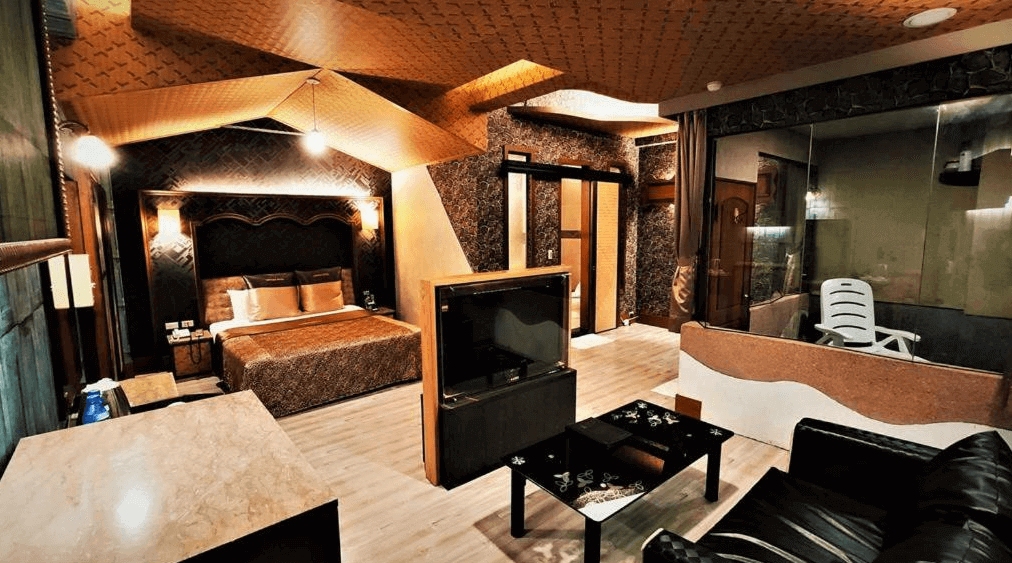 【金山溫泉】金湧泉溫泉汽車旅館的豪華主題房，設計風格大膽且奢華，使用深色系牆面與金銅色調裝飾。房內配有大床、皮革沙發與中間可旋轉的電視，營造高端住宿體驗。玻璃隔間後方設有私人泡湯區，提供隱密的放鬆空間。