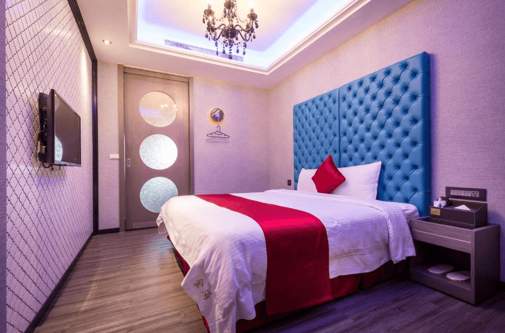 【金山溫泉】金湯溫泉會館的客房，配有藍色立體絎縫床頭板與水晶吊燈，牆面採用柔和淺色調，營造溫馨氛圍。床上鋪有紅色床巾與抱枕，增添質感。房內配備平面電視與簡約衣架，空間設計簡潔且實用。