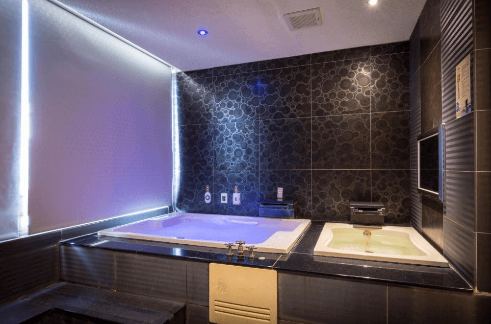 【金山溫泉】金湯溫泉會館的豪華溫泉浴室，配備雙人按摩浴缸與小型溫泉池，提供熱水與冷水選擇。牆面採用黑色拼貼瓷磚，搭配藍紫色燈光，營造現代感十足的泡湯空間。浴室內還設有壁掛式電視，增添休閒娛樂體驗。