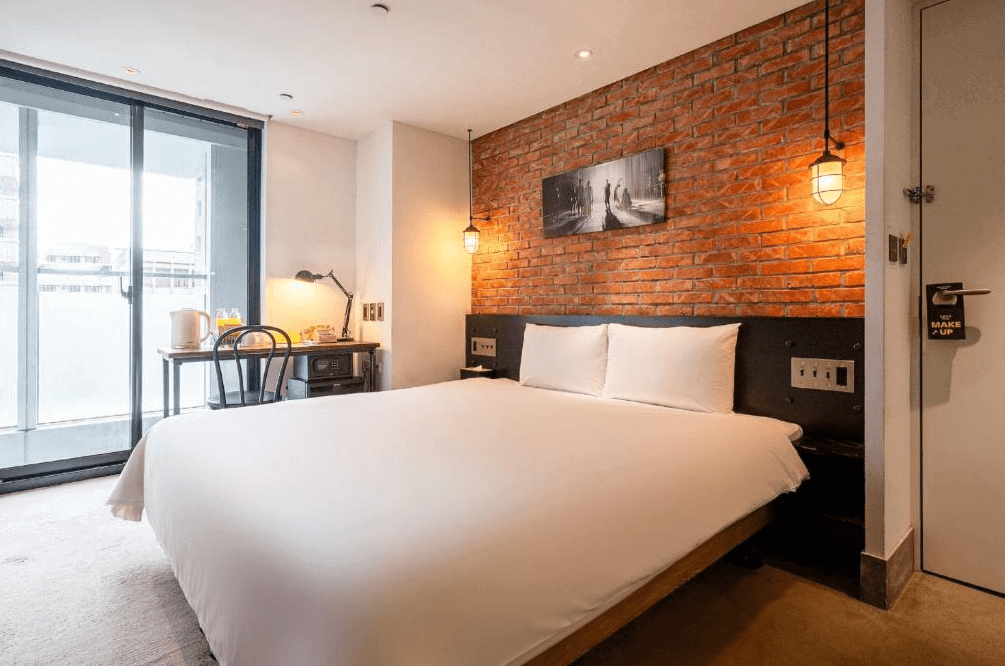 【松山機場住宿】「雀客旅館 台北松江」客房內部，設計風格為工業復古風，牆面以紅磚點綴，搭配溫暖燈光與黑色鐵件裝飾。房內設有一張大床、簡約書桌與玻璃落地窗，提供明亮的採光與舒適的住宿體驗。