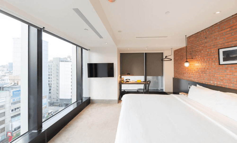 【松山機場住宿】「雀客旅館 台北松江」轉角景觀房，擁有大面積落地窗，提供開闊的市景視野。房內設有一張大床，牆面以紅磚設計搭配黑色金屬燈具，呈現工業風格。房間內部簡約時尚，適合喜愛現代設計的旅客。