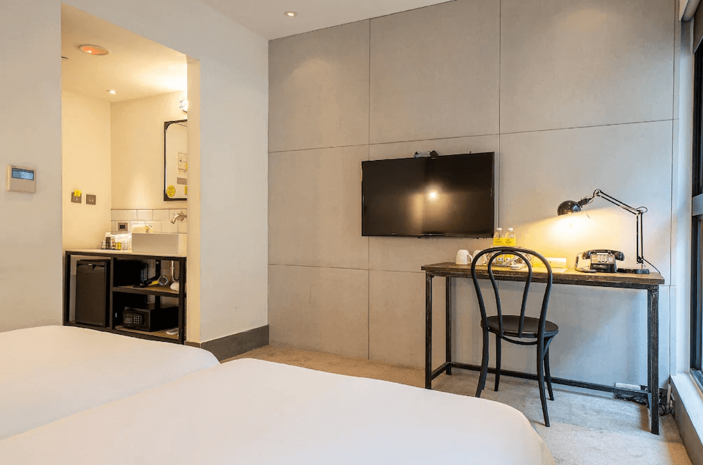 【松山機場住宿】「雀客旅館 台北松江」客房內部，設有雙床配置，簡約工業風設計，牆面採用米色調，搭配黑色金屬與木質家具。房內備有辦公桌、壁掛電視、迷你吧與保險箱，提供實用的住宿體驗。