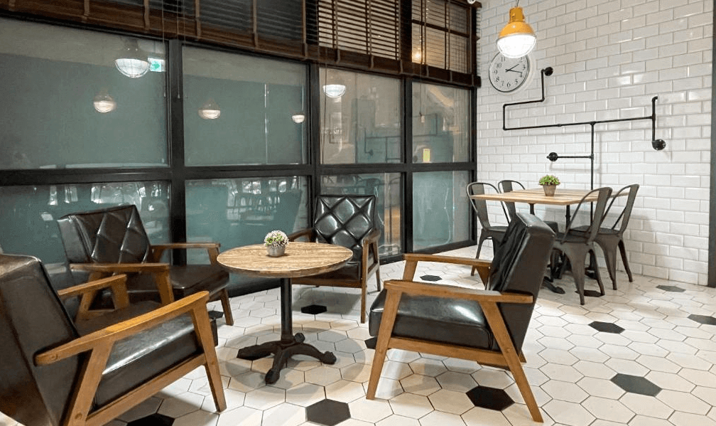 【松山機場住宿】「雀客旅館 台北松江」公共空間，設計融合工業風與北歐簡約風，採用白色磁磚牆面與黑色六角磚地板，搭配復古風皮革座椅與木質桌椅，營造溫馨舒適的社交環境。適合旅客放鬆、交流或簡單辦公。