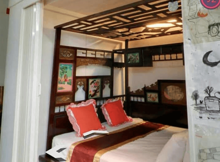 【集集火車站住宿】集集藝術家背包客之家有傳統中式韻味的房間，擁有古色古香的雕花木床與精緻的中式窗花裝飾。床鋪搭配紅色刺繡抱枕與傳統花紋床單，營造典雅的懷舊氛圍，適合喜愛古典風格的旅客入住。