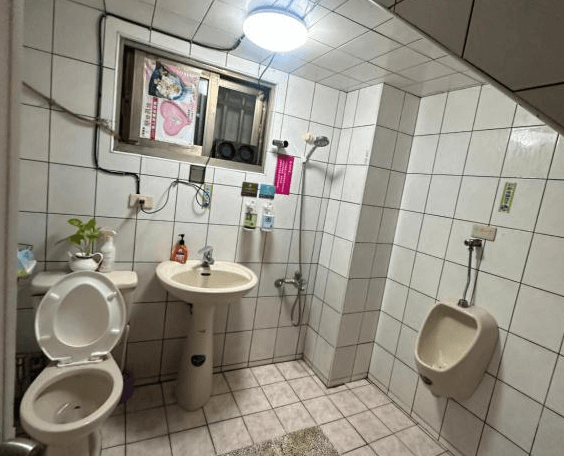 【集集火車站住宿】集集藝術家背包客之家的浴室空間，白色磁磚鋪設的牆面與地板，內含馬桶、洗手台、掛壁式小便斗與簡易花灑淋浴設備。窗戶提供自然通風，並設有簡單的收納架與植栽點綴，使空間更顯整潔實用。