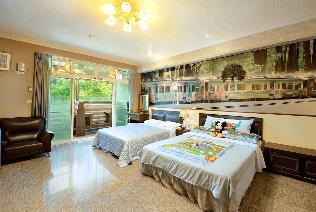 【集集火車站住宿】集集金格民宿的雙床房，牆面裝飾有集集火車站主題的壁畫，增添濃厚的在地特色。一張床鋪上鋪有可愛的兒童床單與米奇玩偶，適合親子住宿。房間內設有皮革沙發、大型落地窗，陽台可俯瞰戶外綠意景色，光線充足。