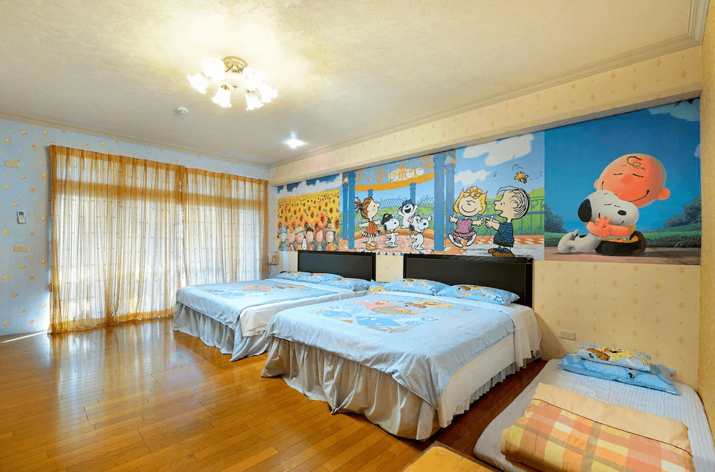 【集集火車站住宿】集集金格民宿可愛童趣風格的四人房，牆上有史努比與《花生漫畫》角色的彩繪壁畫，營造活潑愉悅的氛圍。三張大床舖上淺藍色床單，房內採用木地板與暖色調燈光，搭配柔和的窗簾，使整體空間更加溫馨。