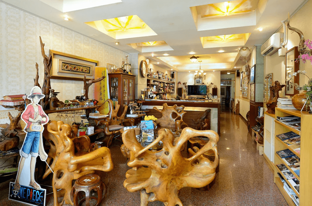 【集集火車站住宿】集集金格民宿充滿木質風格的民宿大廳，擺設多張獨特的原木雕刻桌椅，展現濃厚的自然氣息。大廳內還擺放許多動漫公仔收藏，牆上掛有懷舊裝飾畫與藝術擺件，結合傳統與現代元素，為旅客提供別具特色的公共空間。