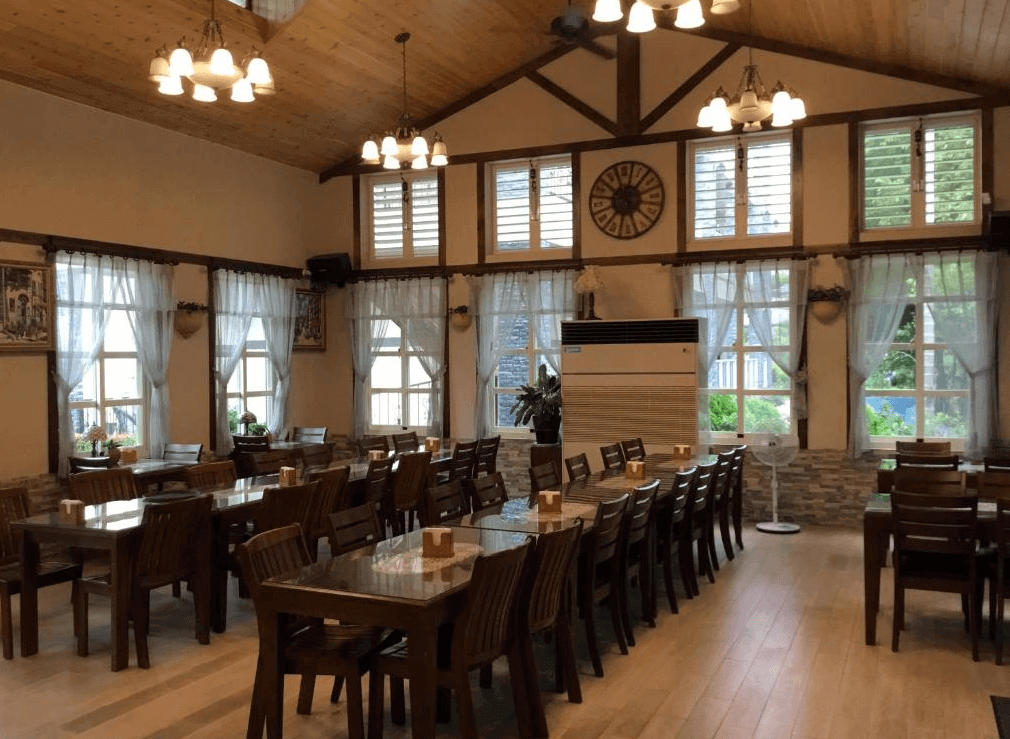 【集集火車站住宿】香榭雅筑民宿的鄉村風格用餐空間，挑高木質屋頂搭配大面積窗戶，讓室內光線明亮柔和。多張木製餐桌與椅子整齊排列，搭配白色窗簾與簡約吊燈，營造溫馨而典雅的用餐環境，適合家庭或團體聚餐。