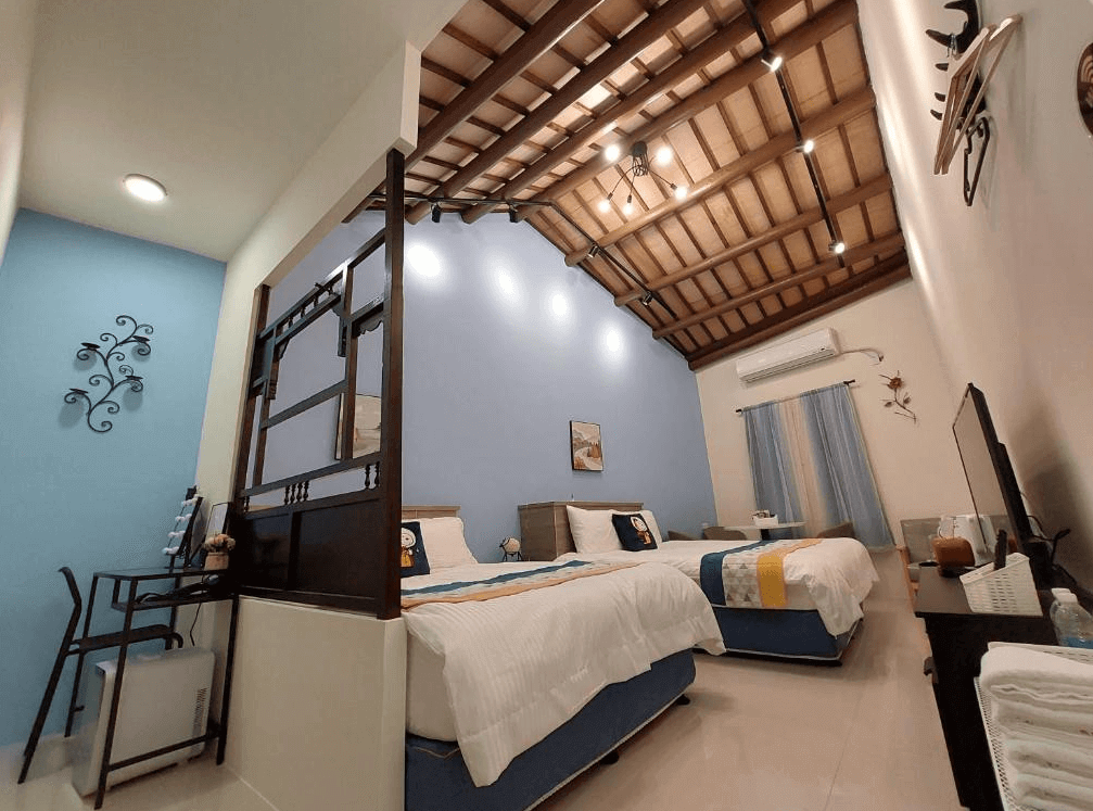 【鹿港住宿】鹿比民宿明亮藍色牆面的雙床房，這間房間的特色在於其開放感和高挑天花板設計。木質梁架的天花板與藍色牆面相得益彰，創造了一種溫暖且有質感的氛圍。房內有兩張床和舒適的沙發，提供住客放鬆的空間。