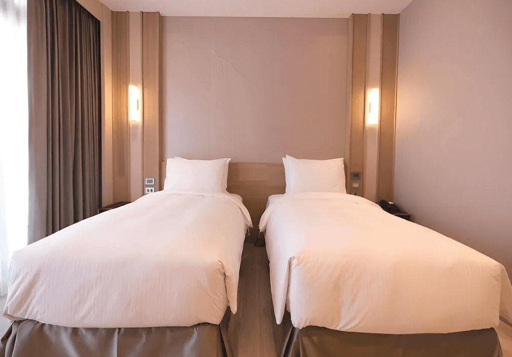 【鹿港住宿】鹿港澄悅酒店的雙床房，這間房間的設計同樣現代且簡約。牆面與窗簾的色調搭配和床頭的簡單裝飾，使整體空間看起來明亮且寬敞。床邊的家具配置實用，提供了足夠的空間來安放個人物品。