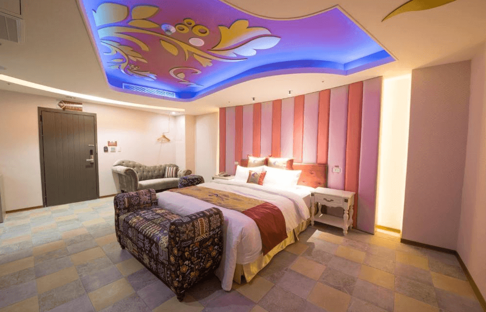 【金山溫泉】鼎帥溫泉旅館的豪華主題房，天花板裝飾著藍色燈光與花卉圖案，增添浪漫氣息。大床搭配粉色與金色的床頭設計，營造華麗氛圍。房內擺放典雅的沙發與長椅，地板鋪設拼接瓷磚，呈現獨特風格。