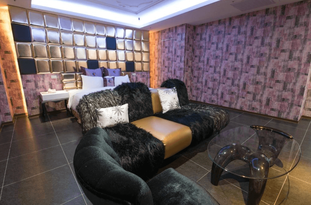 【金山溫泉】鼎帥溫泉旅館摩登風格的豪華客房，搭配金屬質感的床頭牆面與黑色毛絨沙發，營造高端奢華感。房內設有玻璃茶几與柔和燈光，整體色調以金色、黑色與紫色為主，塑造獨特而大膽的設計風格。