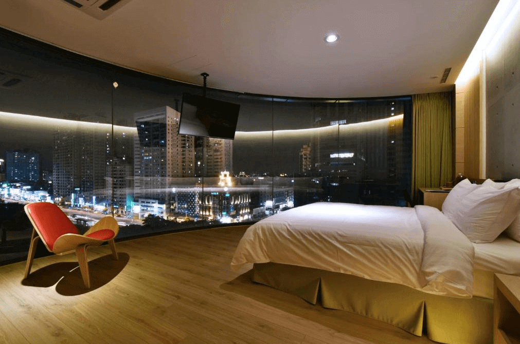 【高雄親子飯店】R14 巨蛋旅店的夜景景觀雙人房，房間擁有大面積的弧形落地窗，可俯瞰高雄市區夜景。室內設計採用木質地板與暖色調燈光，搭配一張舒適的大床與紅色設計感單椅，營造時尚且溫馨的住宿氛圍。