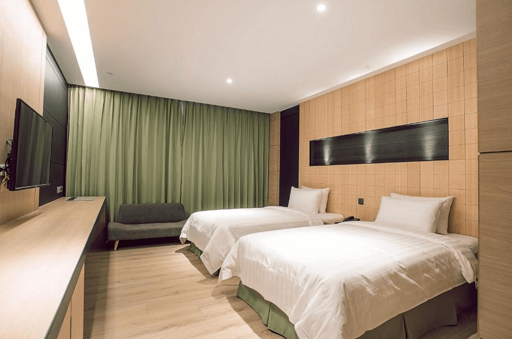 【高雄親子飯店】R14 巨蛋旅店的雙床房，房內採用溫暖的木質調與綠色窗簾，展現沉穩舒適的設計風格。牆面搭配淺色格紋裝飾，並設有簡約的沙發座椅與長型書桌，提供良好的休憩與工作空間。
