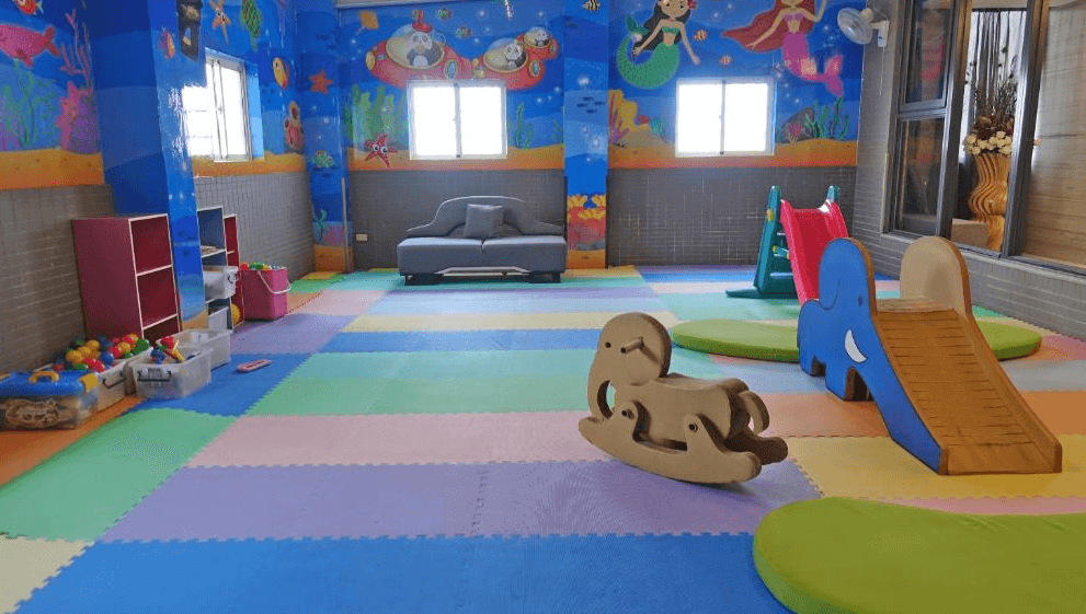【斗六住宿】三好國際酒店的兒童遊戲室，牆面為海底世界主題彩繪，鋪有彩色地墊，設有溜滑梯、搖搖馬與各式積木玩具，中央配有灰色沙發，空間寬敞明亮，適合親子同樂。