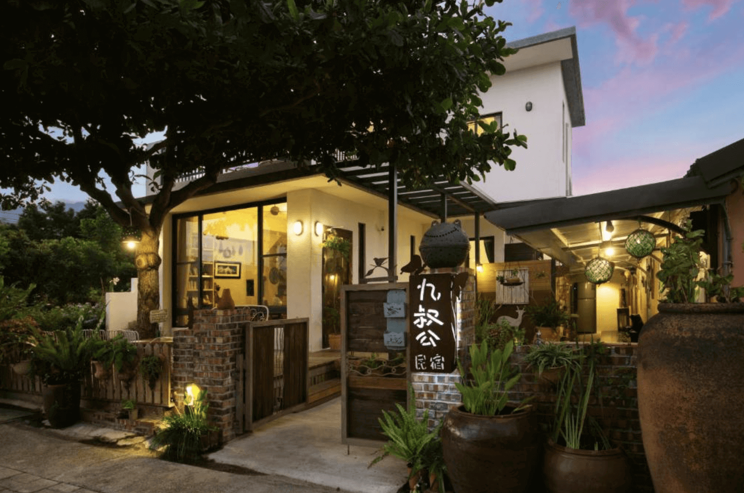 【鹿野住宿】傍晚時分的「九叔公民宿」，整體建築為現代簡約風格，外圍設有綠植與磚砌圍牆，營造溫馨鄉村風情。門口掛有民宿木製招牌，燈光映照下顯得溫暖宜人。庭院內擺放陶甕與各式裝飾，充滿懷舊感，適合喜愛靜謐環境的旅人。