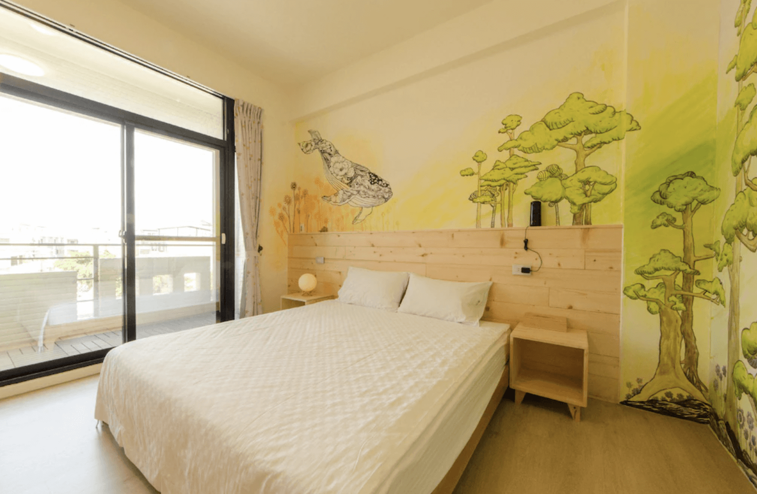 【台南安平住宿】倆倆親子民宿的房間牆面以手繪風格描繪鯨魚與森林景色，色調溫暖自然，床頭板採原木設計，展現出童話般的療癒氛圍。大片落地窗引入自然光線，通往陽台的門設計讓空間更顯通透開闊。