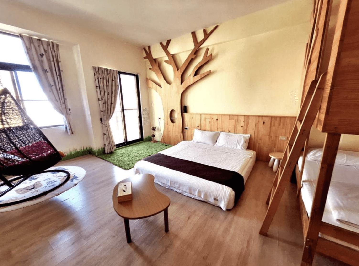 【台南安平住宿】倆倆親子民宿結合樹屋元素與上下舖設計的親子房，牆面裝有立體木質大樹裝飾，地板鋪設綠色草皮地毯，增添森林氛圍。房內設有一張雙人床與上下舖、吊椅與圓桌，空間寬敞，充滿童趣與自然感。