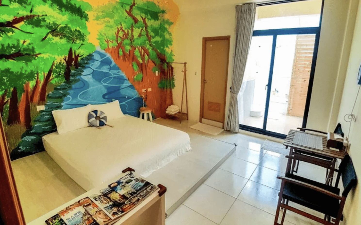 【台南安平住宿】倆倆親子民宿的牆面彩繪蜿蜒河流與濃密樹林，充滿自然生命力，床鋪簡潔俐落，並搭配救生圈造型抱枕點綴。一側設有木質小書桌與衣架，整體氛圍明亮放鬆，適合親子同住。