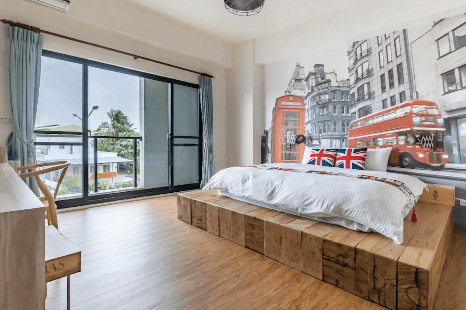 【台南安平住宿】台南搖滾狂民宿英國倫敦為主題的房型，牆面大幅壁畫描繪雙層巴士與紅色電話亭，搭配英國國旗圖案抱枕及原木高架平台床，營造出濃厚的英倫都會氛圍。大片落地窗讓房間採光明亮，並可遠眺窗外綠意街景。