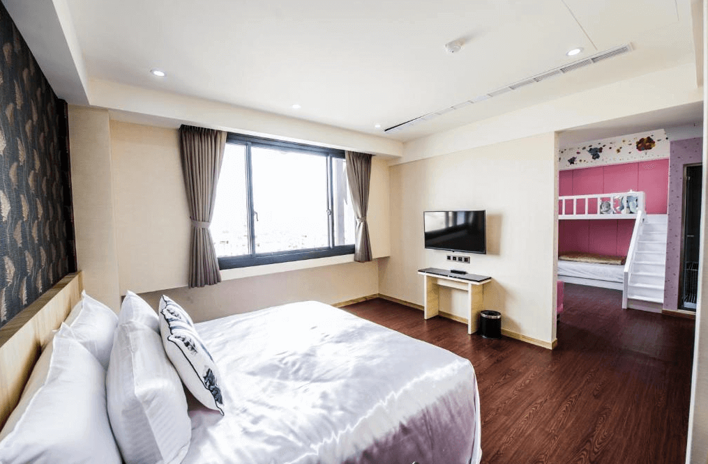 【員林住宿】"彰化飯店"員林紅樓商旅的飯店客房內部，一張大床鋪有白色床單，旁邊有一面大窗戶，增強室內採光。房間內設有壁掛式電視和簡約的書桌。房間旁邊連接一個兒童專屬的遊戲與睡眠區，帶有粉色系設計的上下舖床位，適合親子入住。