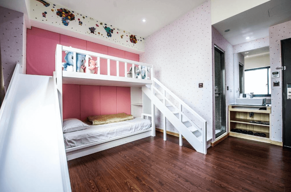 【員林住宿】"彰化飯店"員林紅樓商旅的兒童房間區域，擺放一組粉紅色上下舖床，床上裝飾可愛的絨毛玩偶，並附設一個白色溜滑梯，充滿童趣的設計。旁邊是乾濕分離的衛浴設備，整體空間明亮寬敞。