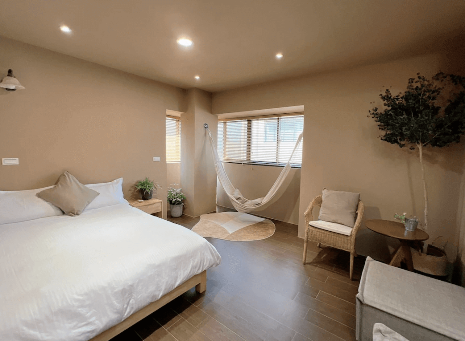 【台南安平住宿】四季洋房的房間採用溫潤的大地色系設計，搭配簡約木質家具與植栽點綴，展現出舒適溫馨的自然風格。一角設有吊床與小圓地毯，適合放鬆閱讀或小憩，整體空間寧靜療癒，氛圍溫柔宜人。