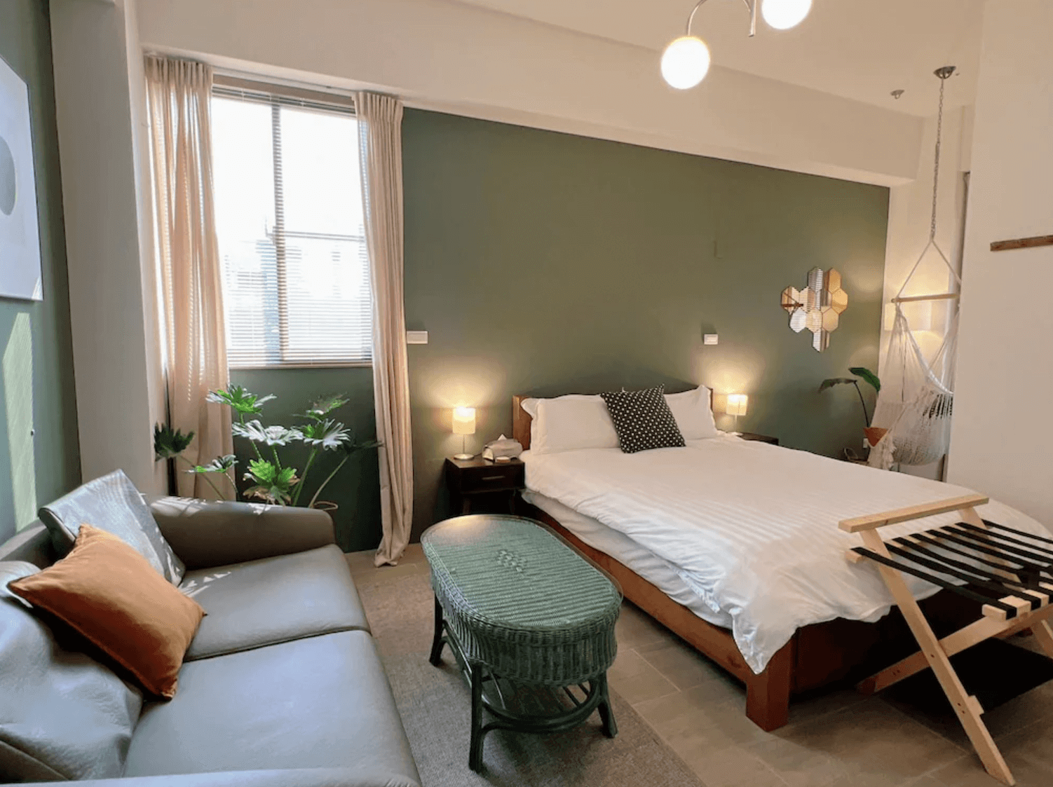 【台南安平住宿】四季洋房的房內以墨綠色主牆為視覺重點，搭配淺木家具與柔和燈光，營造出現代且溫暖的居家感。配置舒適雙人床、皮沙發與吊床椅，提供放鬆自在的住宿體驗，適合情侶或小家庭入住。