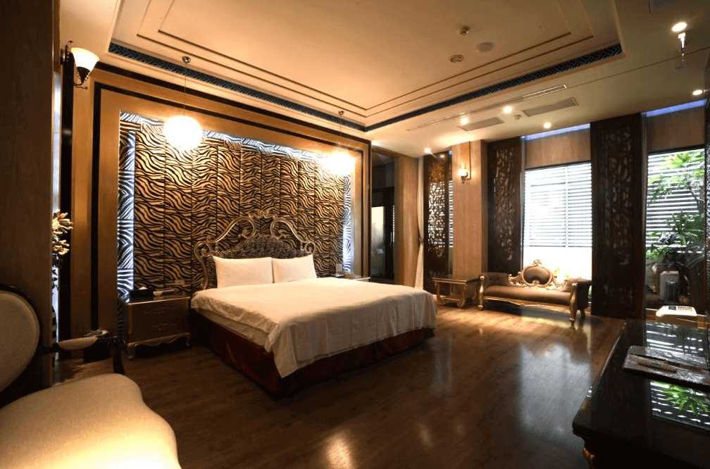 【斗六住宿】天上人間精品旅館的雙人套房，房內設有大型雙人床，床頭為立體浮雕動物紋設計，整體空間以金色與深木色為主，搭配雕花家具和典雅吊燈，窗邊有古典風格沙發與落地窗，營造出寬敞舒適且氣派的氛圍。