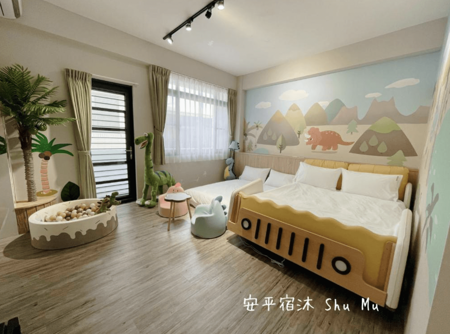 【台南安平住宿】安平宿沐的親子房設有兩張大床，其中一張為造型童趣的越野車床。牆面繪有可愛的恐龍與山景插畫，房內擺放恐龍玩偶、溜滑梯造型小椅與球池，營造出活潑有趣的遊戲氛圍，適合親子共度歡樂時光。