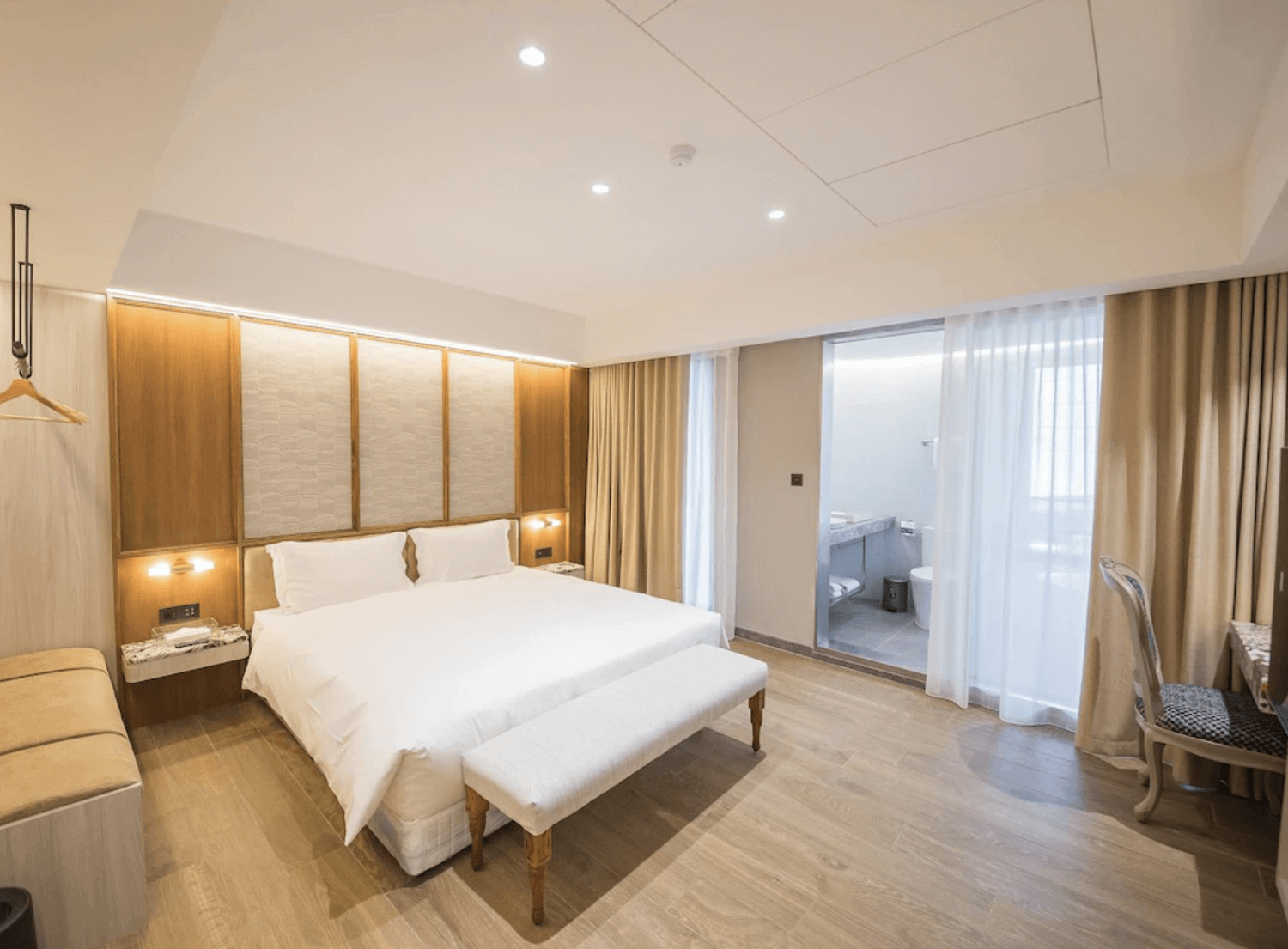 【台南安平住宿】安平留飯店的雙人房一景，配有一張大型雙人床、沙發長椅及通往衛浴的開放式通道，房內採暖色燈光與木地板鋪設，氣氛溫馨且舒適。