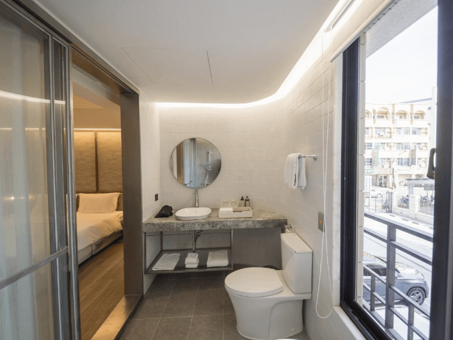 【台南安平住宿】安平留飯店的衛浴空間，採用白色磁磚與圓形鏡面設計，搭配大面窗戶引入自然光，洗手台與馬桶分區明確，整體明亮又具現代感。