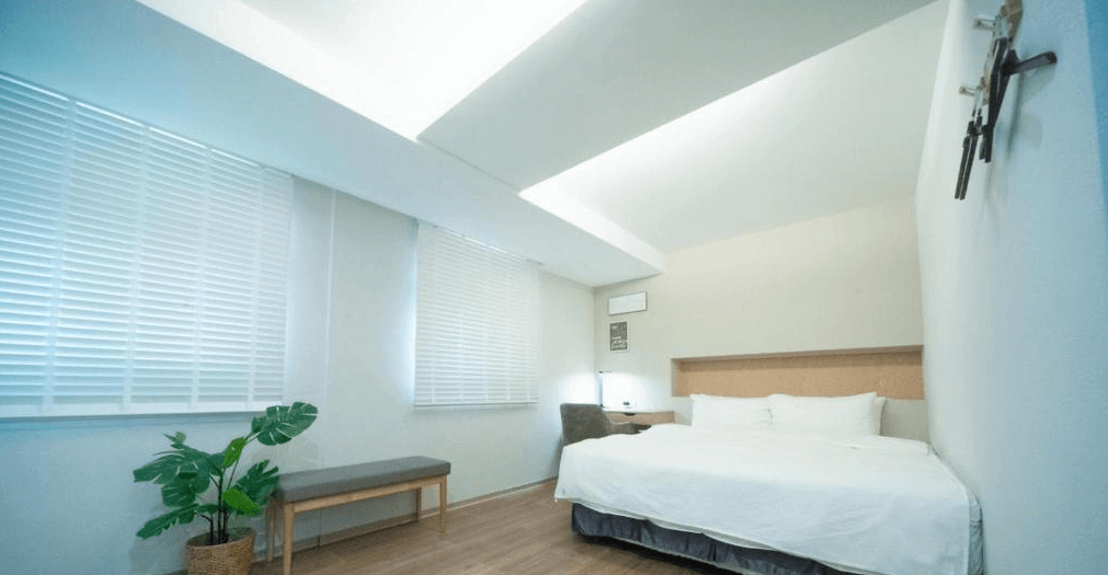 【虎尾住宿】小9窩BNB的簡約現代風格的客房，大片白色百葉窗與木質地板打造清爽氛圍，白色床鋪旁設有書桌椅與小盆栽點綴，空間寬敞明亮，適合悠閒放鬆。