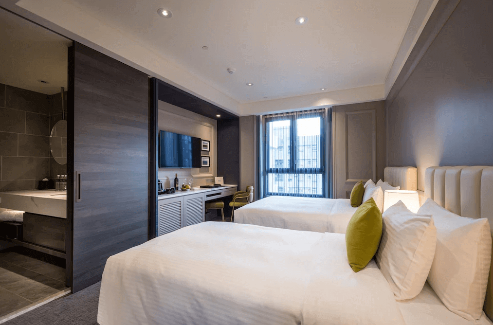 【高雄親子飯店】帕可麗酒店溫馨典雅的雙床房，房內設計簡約大方，以柔和的米色和木質調為主。床鋪擺放整齊，搭配綠色抱枕增添活力感。房內設有落地窗，可引入自然光，並有書桌、電視和迷你吧設備。浴室與房間之間採用木質滑門設計，兼具隱私與空間感。