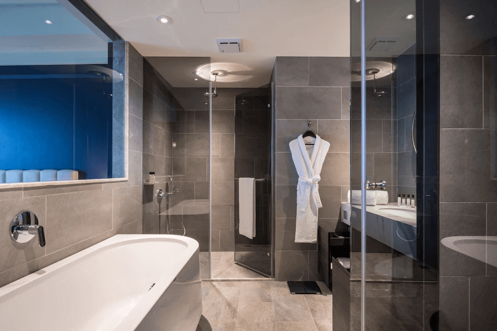 【高雄親子飯店】帕可麗酒店的高級大理石衛浴空間，採用灰色石紋磁磚，呈現時尚摩登氛圍。房內設有寬敞的獨立浴缸與玻璃淋浴間，並備有高級浴袍和品牌盥洗用品。洗手台區域配備大面鏡子與檯面收納，整體設計兼具實用性與高級感。